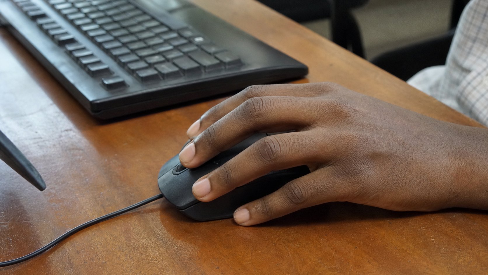 A right hand resting on a computer mouse on a wooden desk.