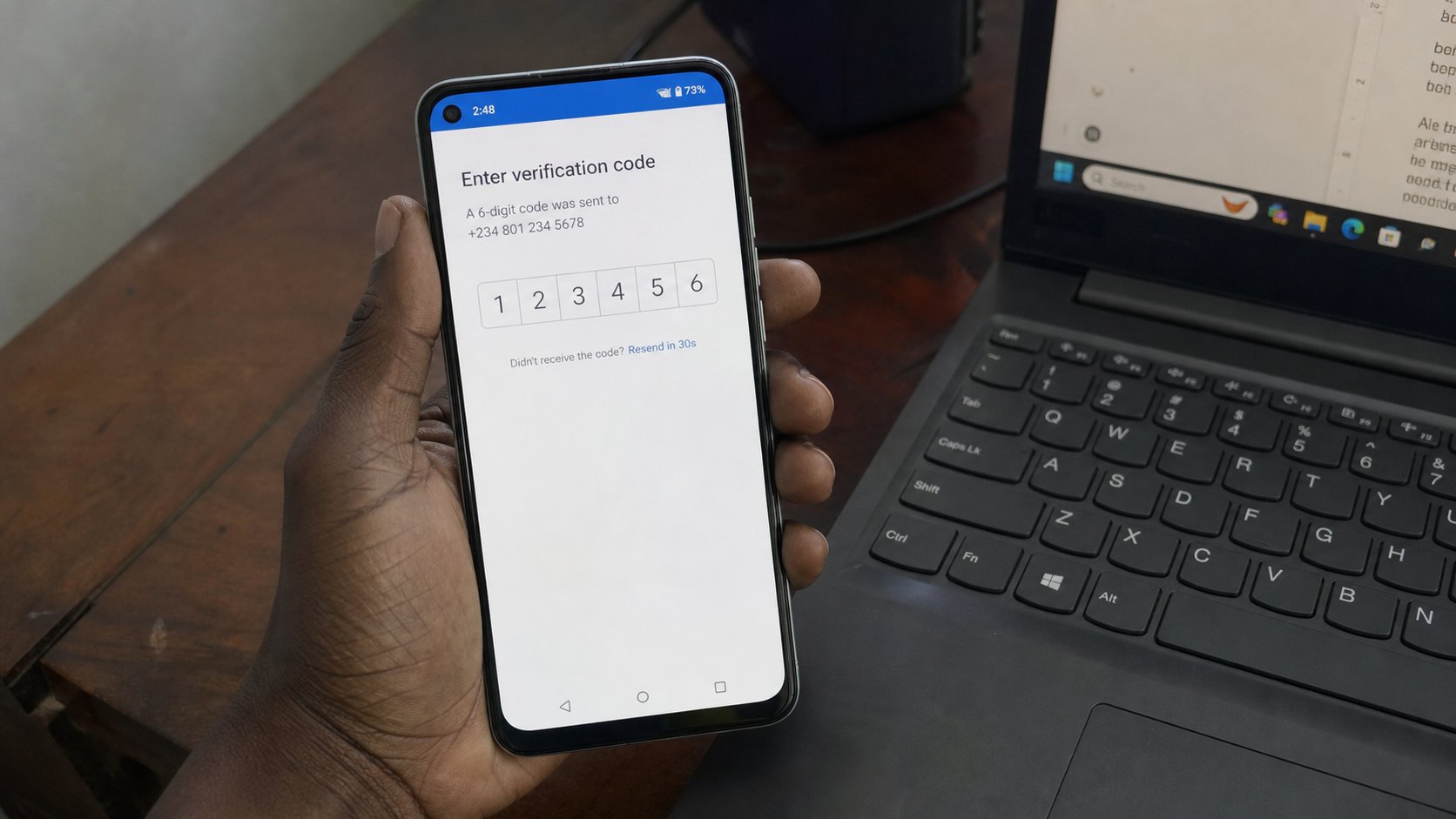 A hand holding a phone that shows a six-digit verification code, laptop open beside it.