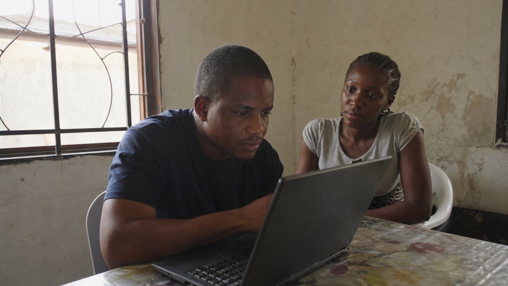 Two people taking turns at one laptop in a modest room.