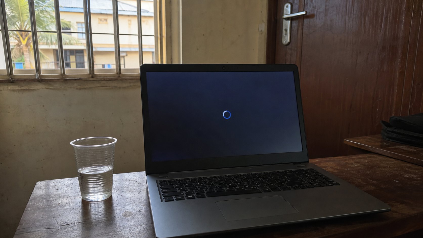 A laptop on a wooden desk with an update in progress, a glass of water beside it.