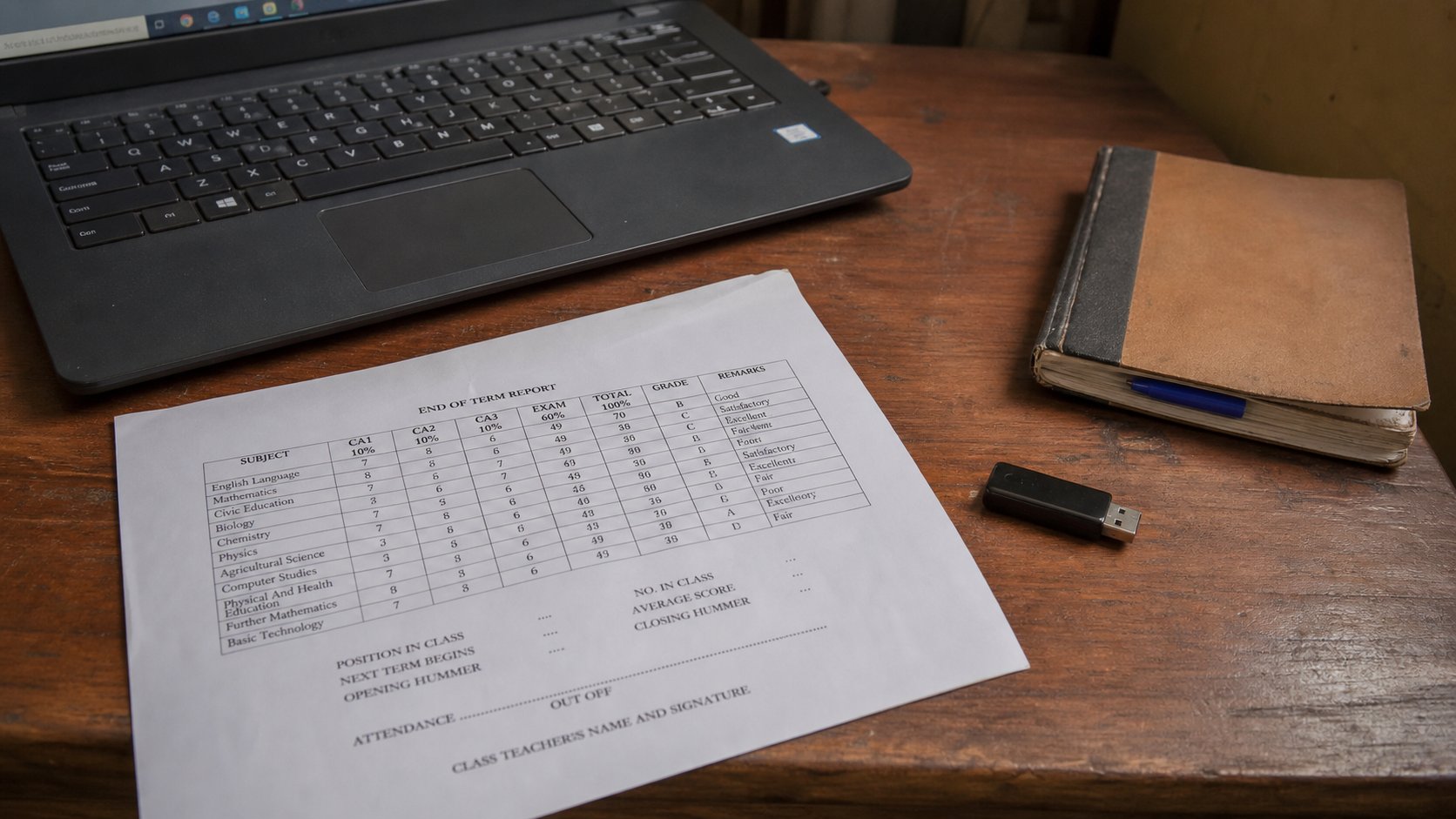 A USB flash drive, a printed document and a notebook on a wooden desk beside a laptop.