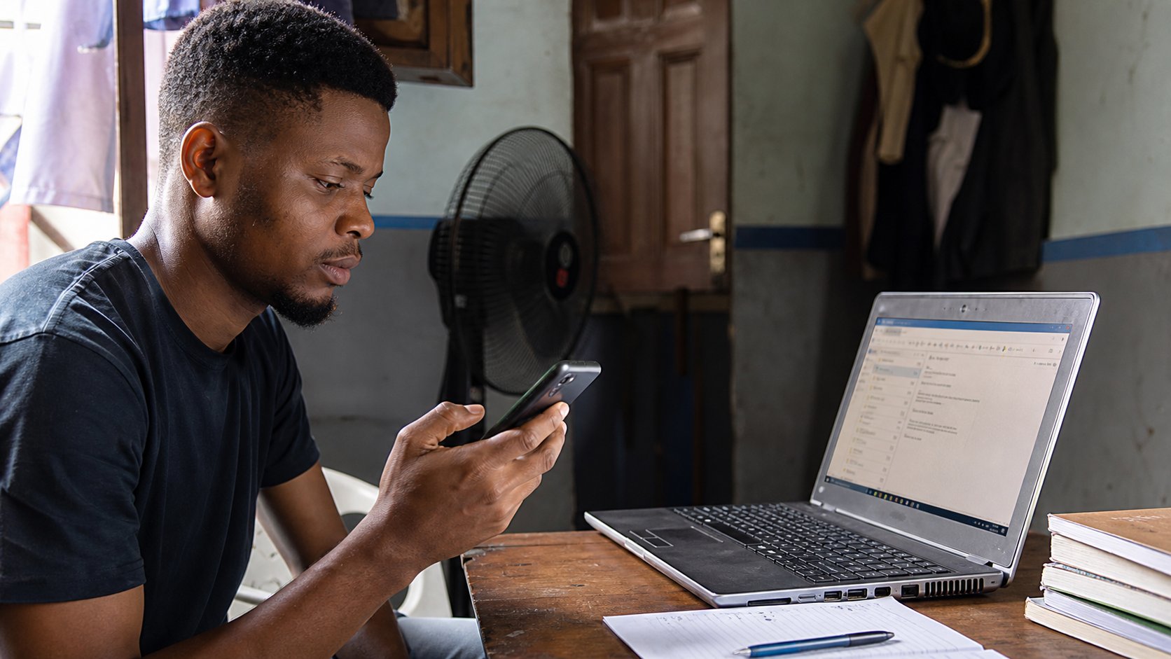 A young man holding a phone while a laptop with email is open on the desk.