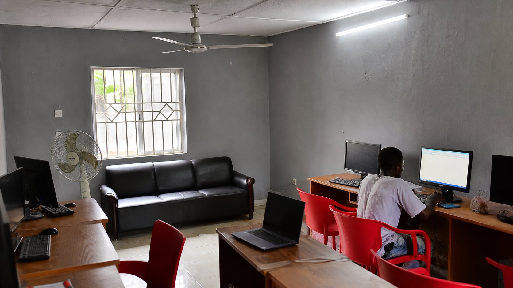 Learners at laptops and desktops in the Ebony Road classroom, standing fan in the foreground.